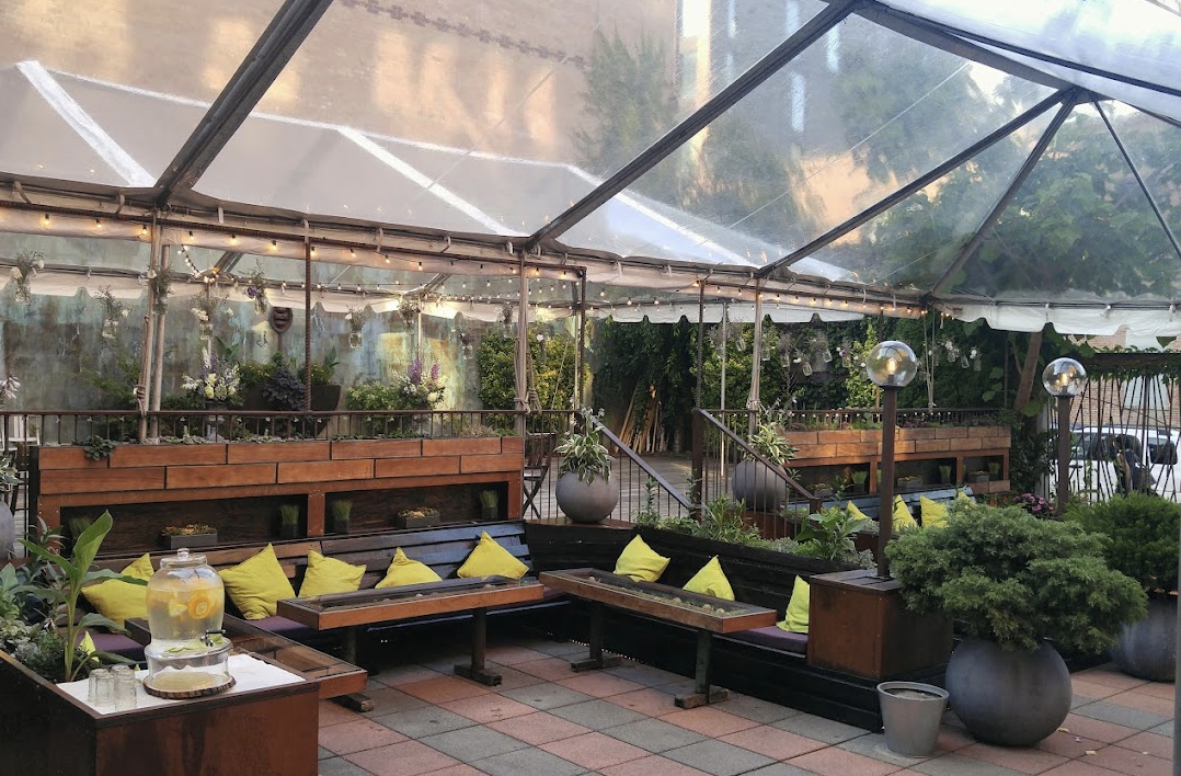 Clear-top tent covering a rooftop lounge with wood benches and yellow cushions